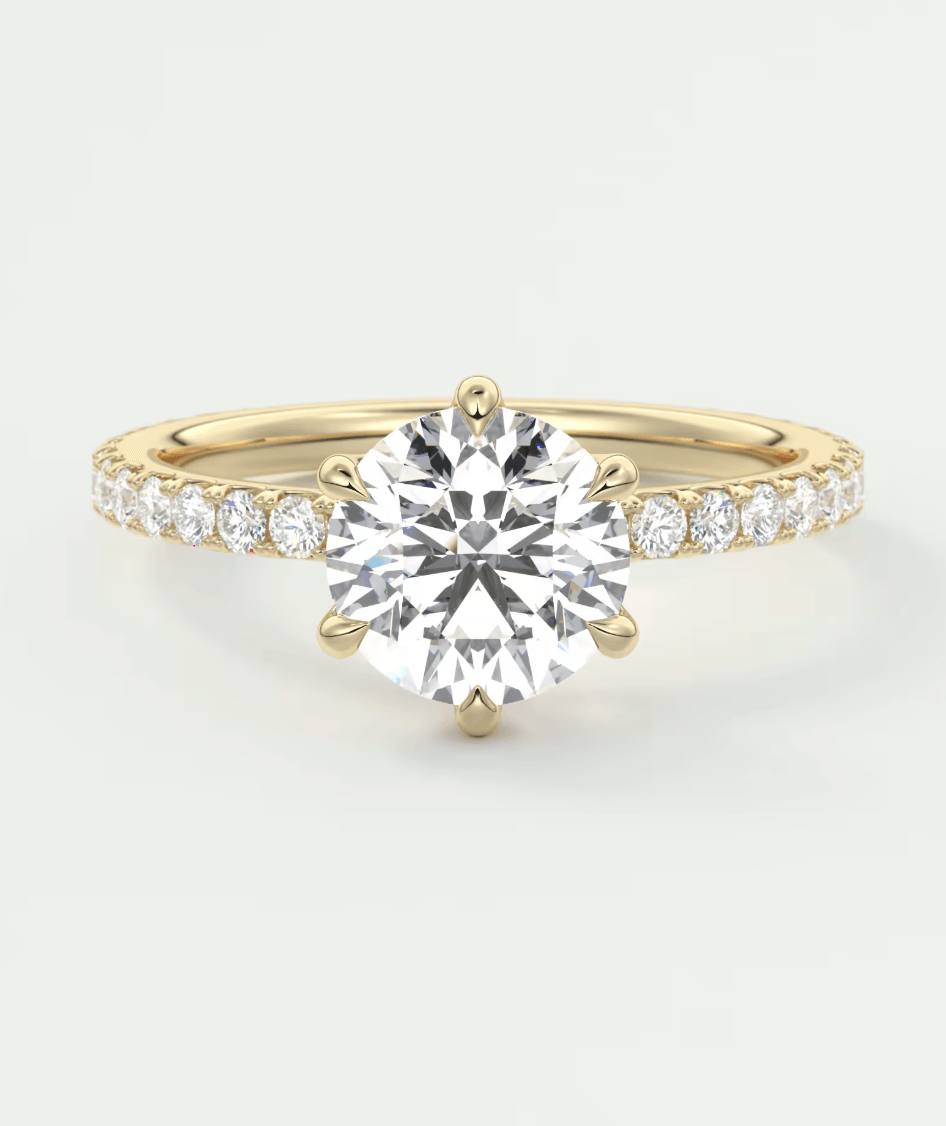 A round-cut diamond or moissanite engagement ring with a pavé band set in a gold metal band, displayed against a white background.