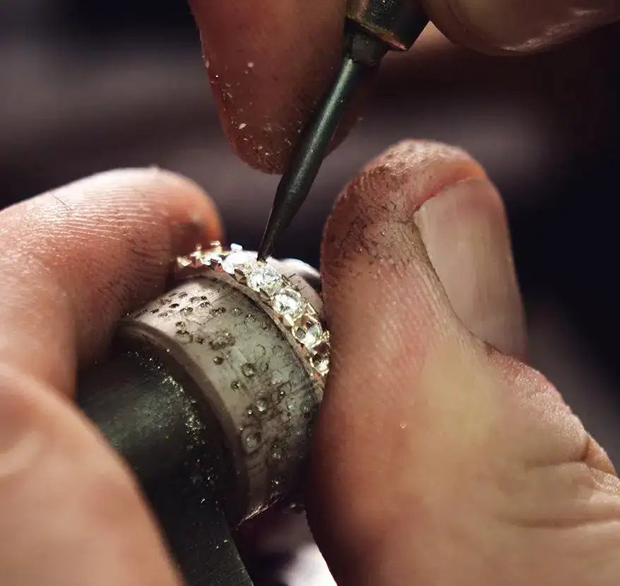 A silver ring with embedded clear gemstones being meticulously crafted by a jeweler’s hands.