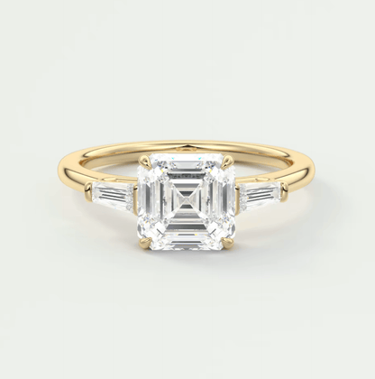 A gold three-stone engagement ring with a central Asscher-cut diamond or moissanite, flanked by two tapered baguette side stones, and a band with pavé diamonds.