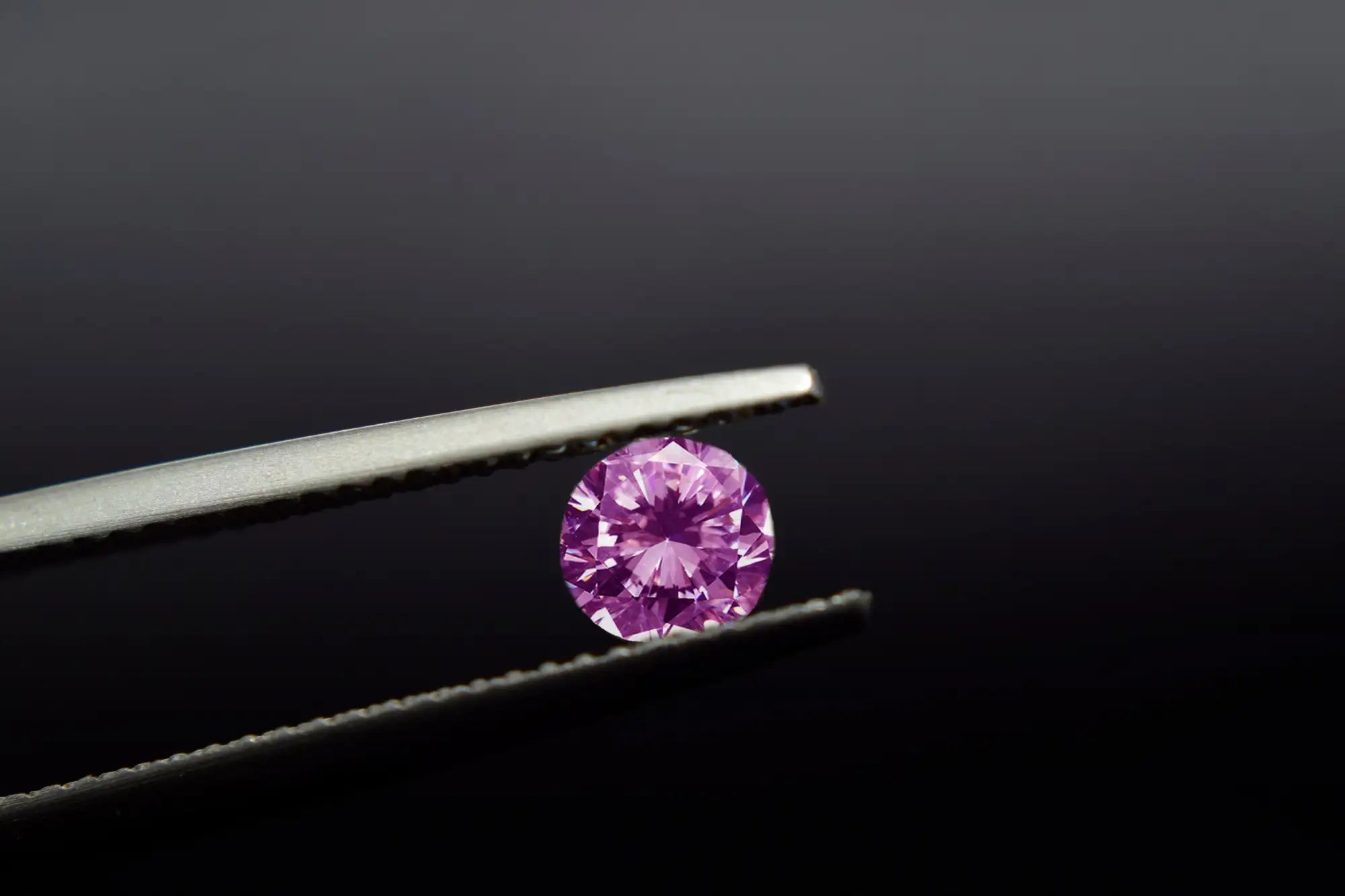 A round, vivid pink diamond held delicately between precision tweezers.
