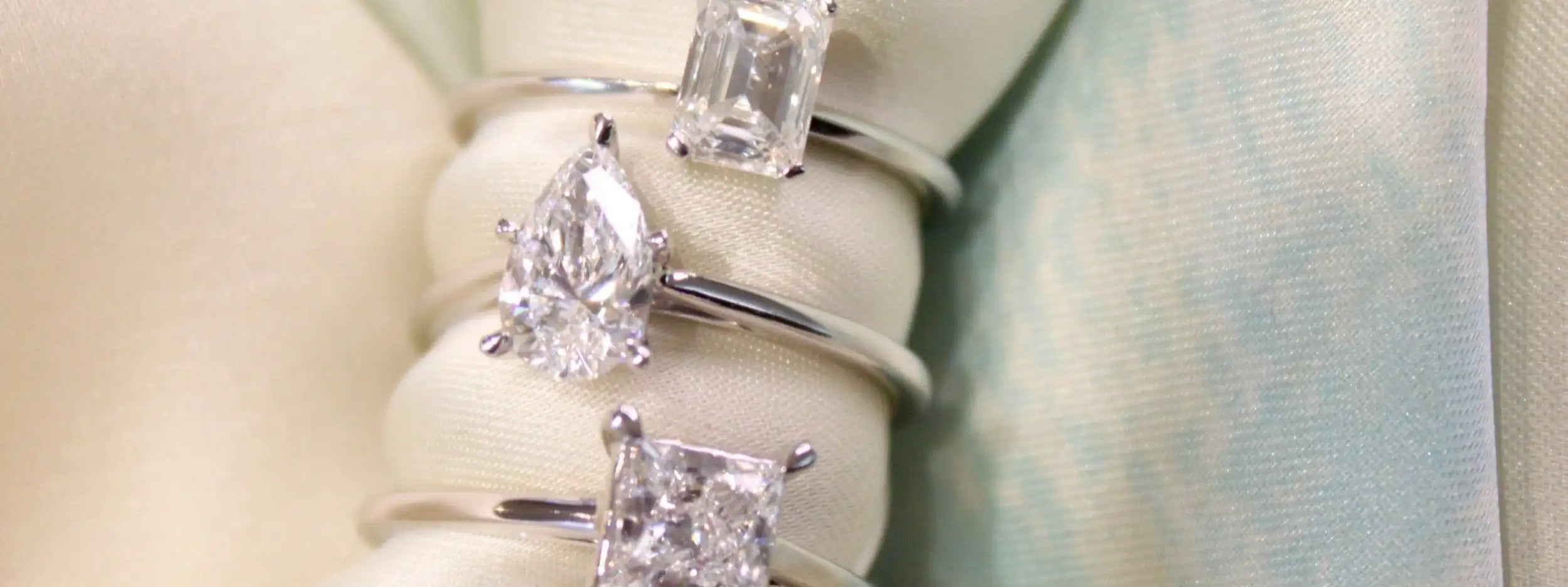 Three elegant diamond rings with varying cuts—pear, emerald, and princess—are displayed on a soft white cushion.